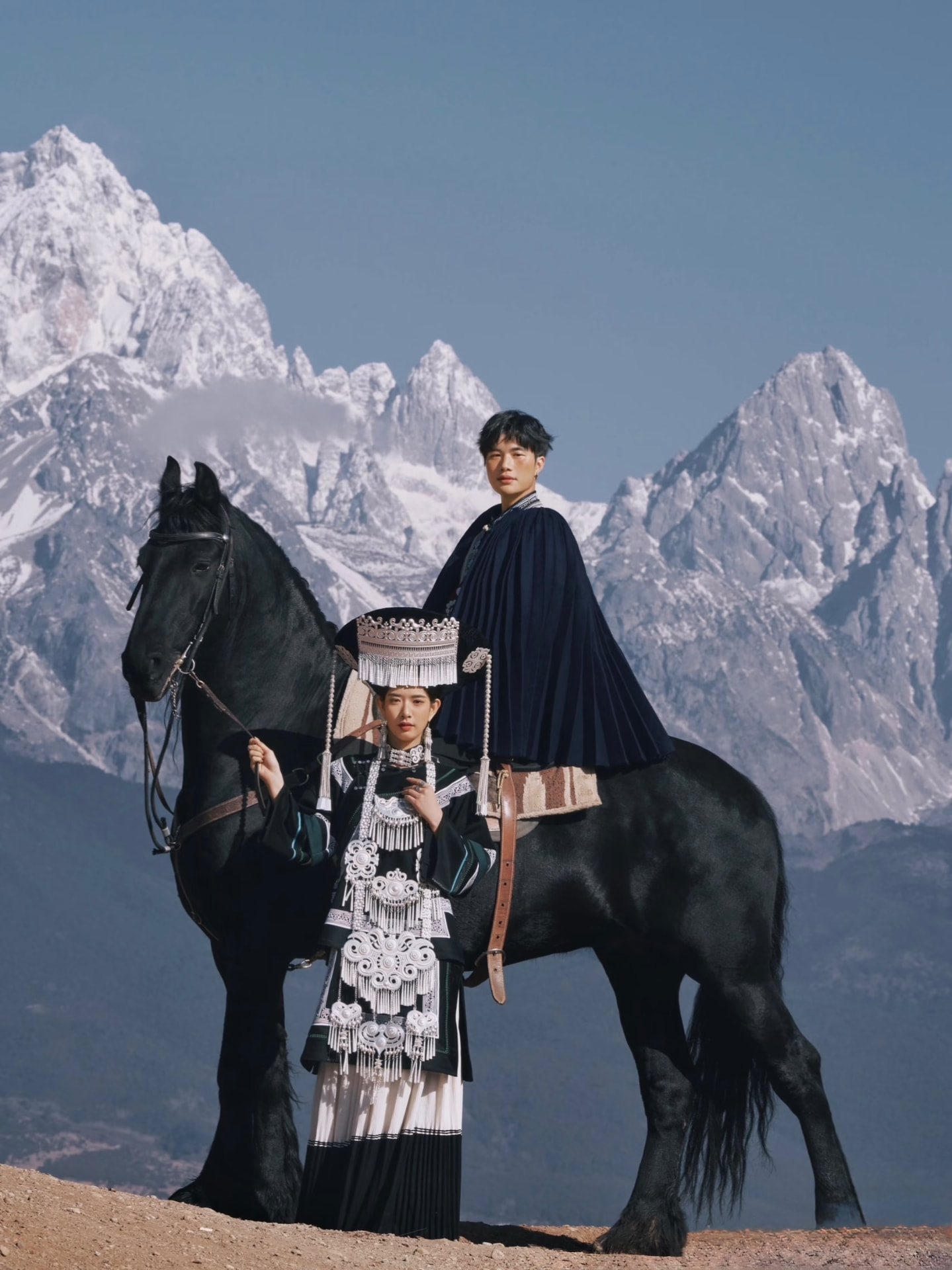 Two people in traditional Yunnan dress with a black horse against Jade Dragon Snow Mountain
